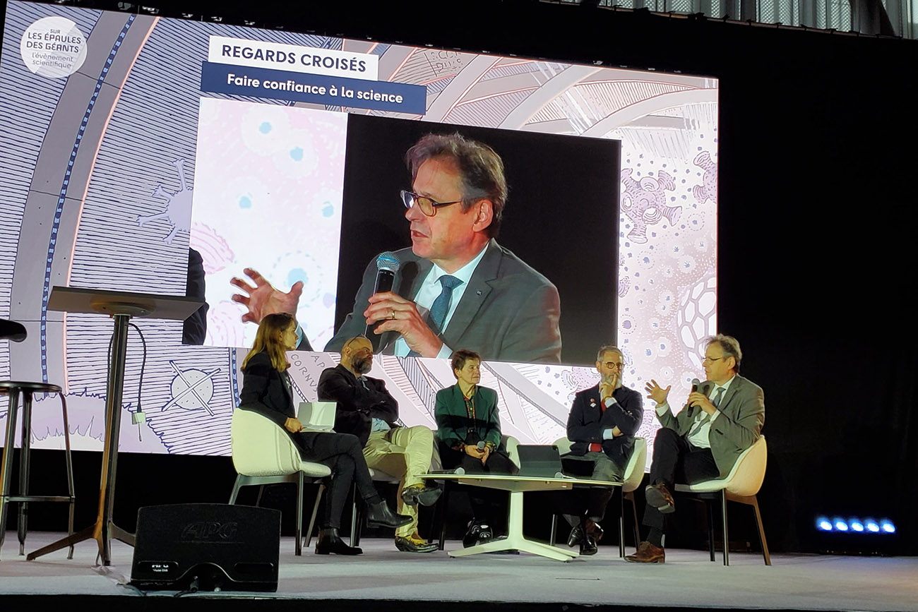 Table ronde avec trois hommes et deux femme, vue du bord de la scène, avec Alain Schuhl tenant le micro reporté sur un grand écran derrière la table ronde