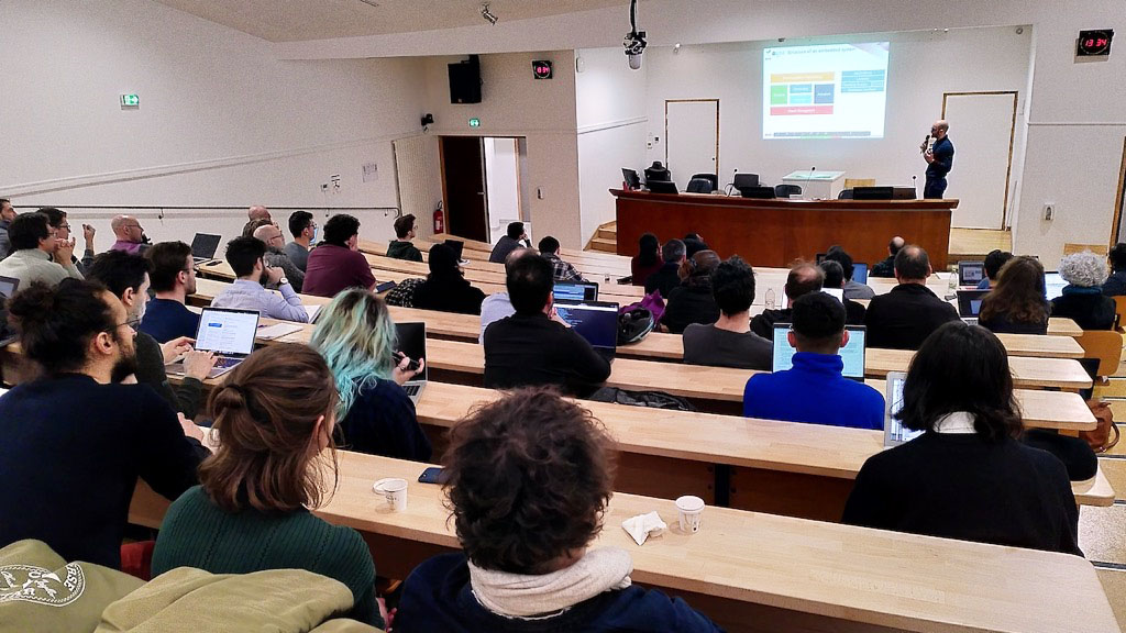 Salle type amphi pleine avec un homme présentant des slides au fond